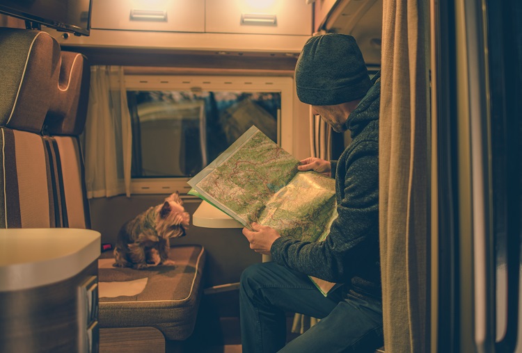 Caucasian Men Planning Trip in His Camper Studying Local Map. His Dog is Seating on Opposite Side of the Motorhome Table. Rving with Pet.
